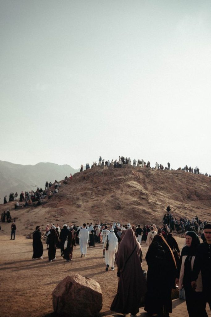 Crowd of pilgrims wearing traditional attire gather at a sandy hill under a clear sky.