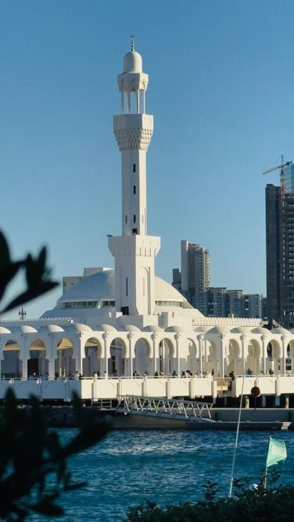 Stunning view of a white mosque with minaret by the coastal waters under a clear blue sky.